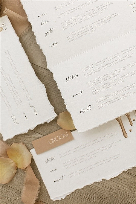 Wedding menus with deckle edge menu card in calligraphy, wax seal and ribbon beside a place card on a wooden tabletop with rose petals