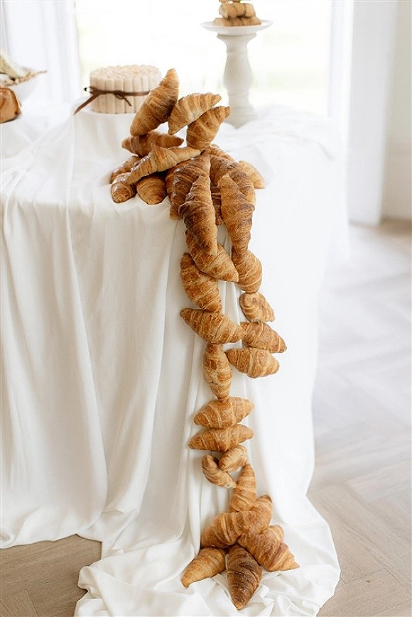 Wedding dessert table with a croissant dessert display on white tablecloth, small cake on stand, ribbon accents, and airy draping in window light