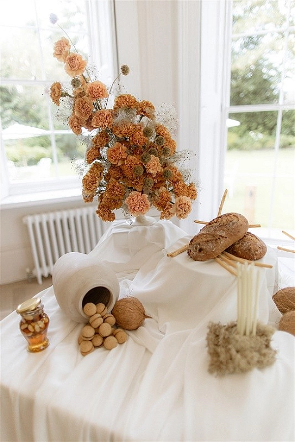 Wedding tablescape with terracotta flowers and a floral centerpiece, taper candles, bread loaves, walnuts, and honey jar by large windows
