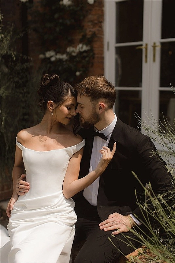 Couple portrait of bride in an off the shoulder wedding dress and groom in tuxedo, touching foreheads by French doors and brick wall greenery