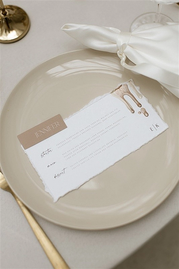 Wedding place setting with a wedding menu card clipped over a dinner plate, ribbon bow detail, gold flatware, and wine glass on linen tablecloth