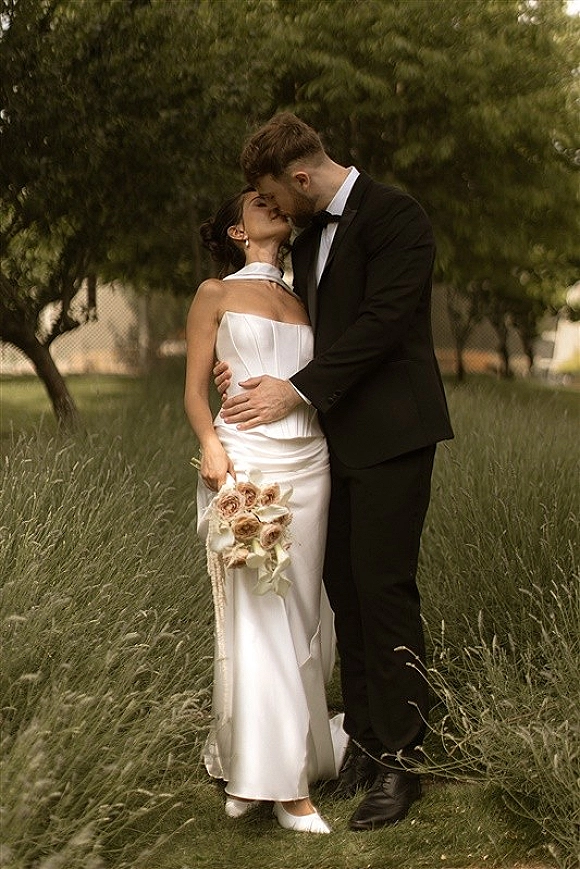 Wedding kiss portrait of bride and groom kissing, she in strapless satin dress holding rose bouquet, in tall grass garden lawn backdrop