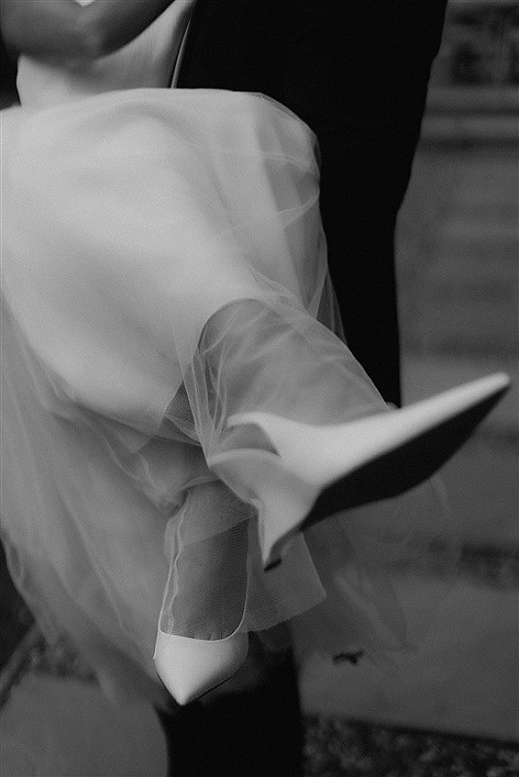 Bridal shoe detail with white pointed toe heels beneath tulle wedding dress fabric as the bride sits on stone steps