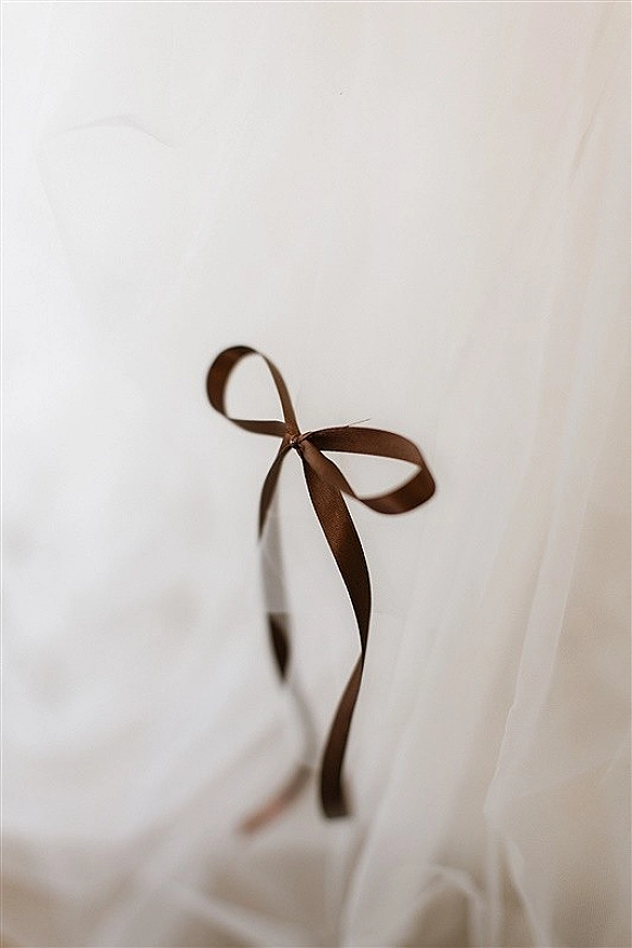 Wedding dress detail with a brown satin ribbon bow tied at the waist over soft tulle layers against a clean white fabric backdrop