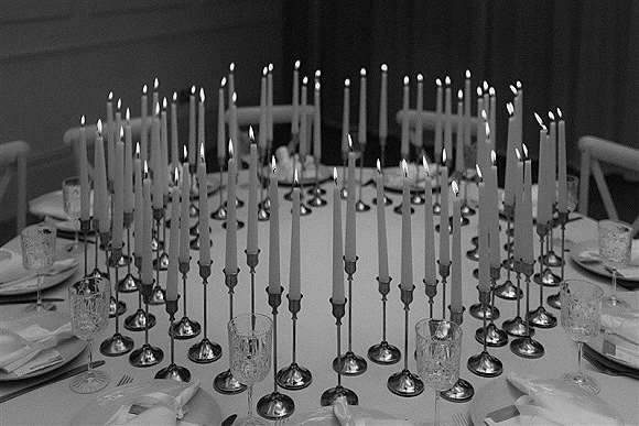 Reception tablescape with a wedding candle centerpiece of white taper candles in silver candlesticks, set on linens with glassware against a dark wall