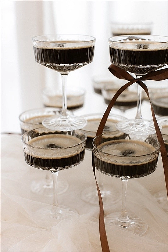 Wedding champagne coupe glasses filled with a dark sparkling drink, crystal stems tied with a satin ribbon on a white tablecloth in soft daylight