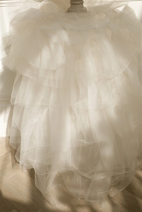 Wedding dress close-up showing a tulle wedding dress with layered ruffle skirt on a hanger by a window, soft light on wooden floor