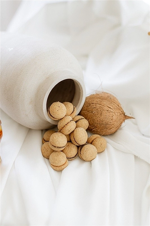 Wedding favors in ceramic jar with mini sandwich cookies and coconut accents, arranged on white fabric over a white draped cloth background