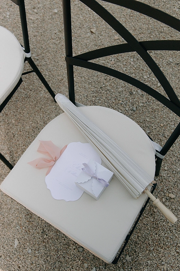 Ceremony chair details with wedding ceremony programs tied in a pastel ribbon bow, favor box and parasol on a cushioned folding chair on gravel