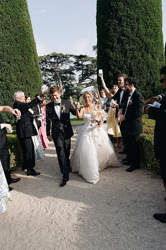 Wedding recessional newlyweds walking aisle as guests toss confetti on a garden pathway lined with evergreens under a blue sky