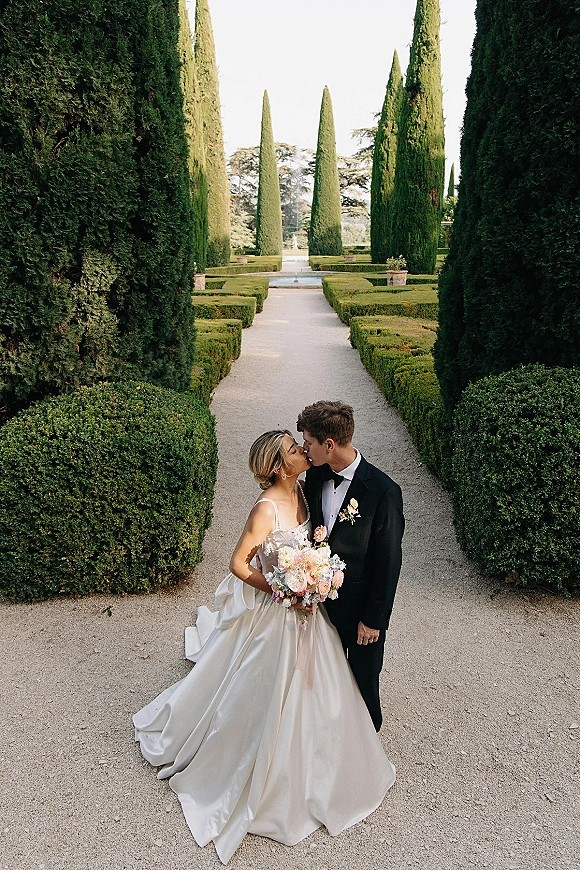 Wedding kiss portrait of bride and groom kissing on a gravel garden path, her bouquet and satin ball gown beside his black tuxedo