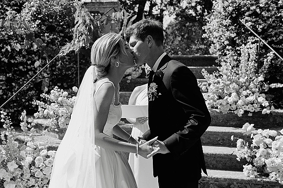 Wedding kiss as bride and groom hold hands on floral-lined stone steps, veil and tuxedo details, officiant behind in garden setting