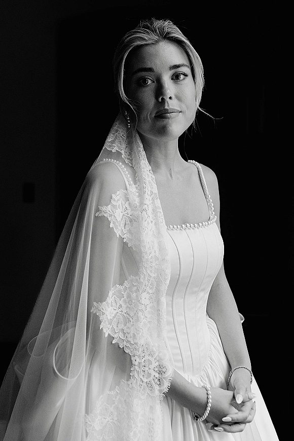 Bridal portrait of a bride in a black and white bridal portrait wearing a lace veil with pearl-trim dress against a dark backdrop