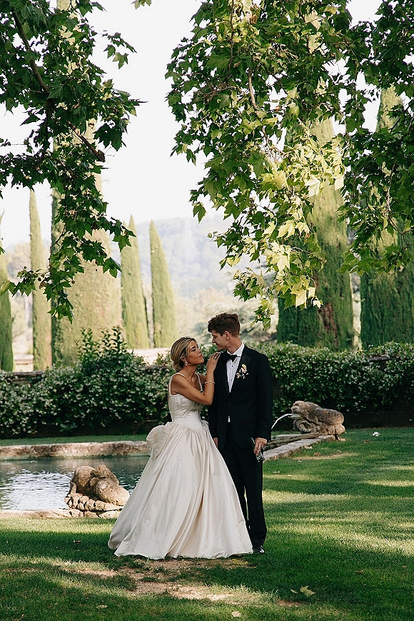Couple portrait of bride and groom portrait as she touches his lapel, strapless gown and tuxedo by a stone fountain in a garden lawn