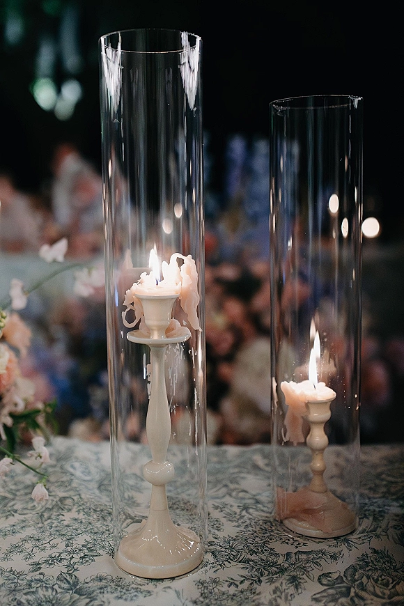 Wedding centerpiece candles in tall hurricane vase candles, with taper wax drips and floral accents on a patterned tablecloth, bokeh reception lights