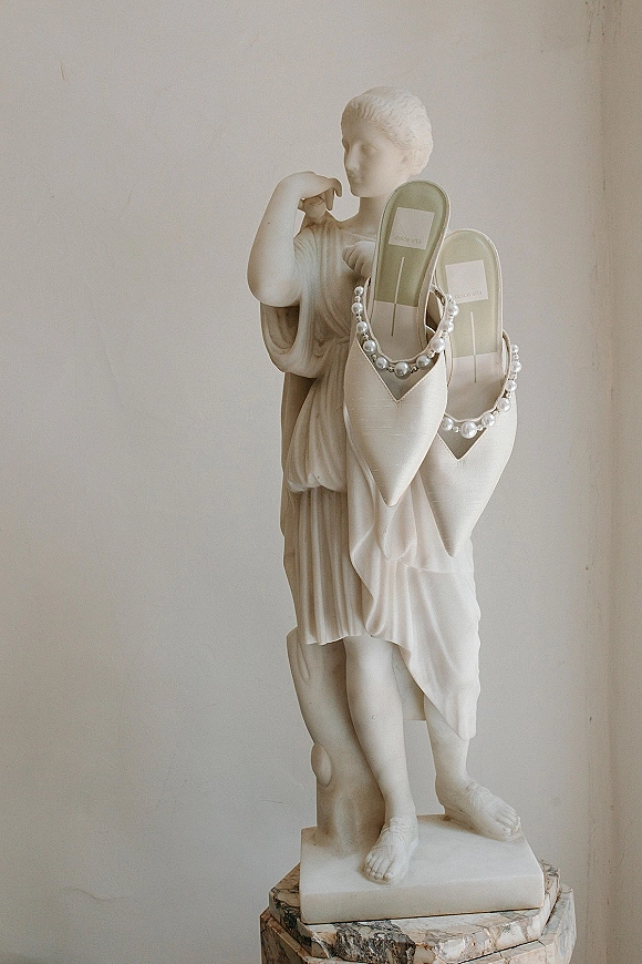 Bridal shoes with pearl wedding shoes straps, white pointed-toe mules displayed on a marble pedestal beside a classical statue in studio setting