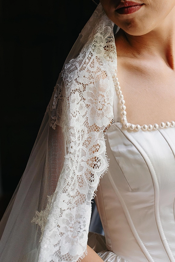Bridal dress details with a lace wedding veil draped over a strapless satin bodice and pearl necklace against a dark backdrop