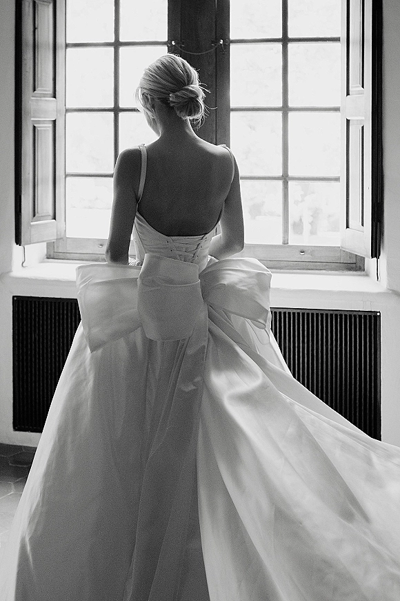 Bridal portrait of a bride by window in a corset-back spaghetti-strap gown with oversized bow and updo, framed by window panes