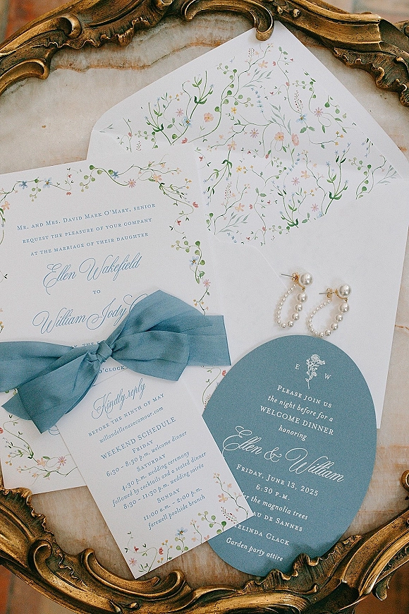 Invitation suite of floral wedding invitations with blue ribbon bow and pearl hoop earrings on marble beside an ornate gold frame