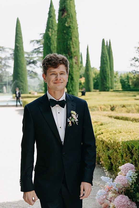 Groom portrait in a black tuxedo with bow tie and boutonniere, standing on a garden path lined with hedges and cypress trees.