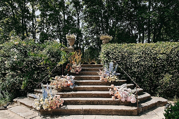 Floral staircase decor with wedding staircase flowers lining stone steps, pastel roses and hydrangeas on iron railings beside hedge walls