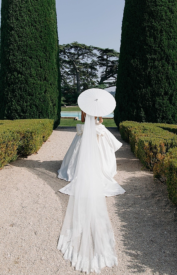 Bridal portrait of a bride from behind in a ball gown with bow, long veil and white parasol on a gravel path in a formal garden