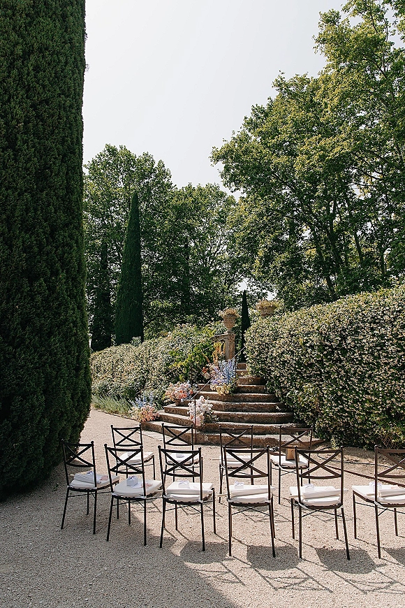 Outdoor ceremony setup with ceremony chairs in rows and white cushions lining stone steps, pastel florals and urn planters in a formal garden setting