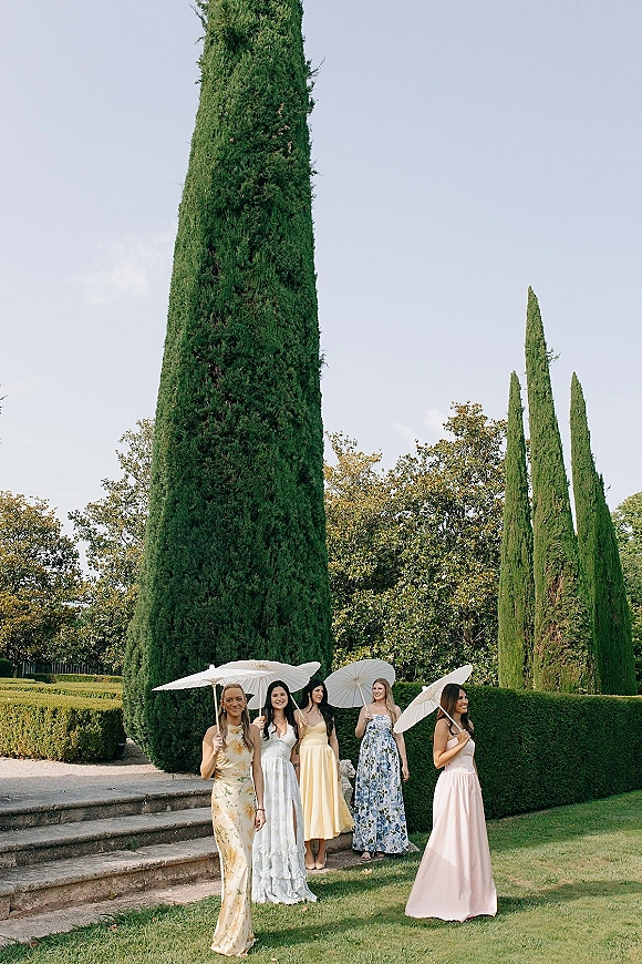 Bridesmaid group photo of pastel-dressed bridesmaids with parasols on stone steps in a garden with cypress trees and blue sky