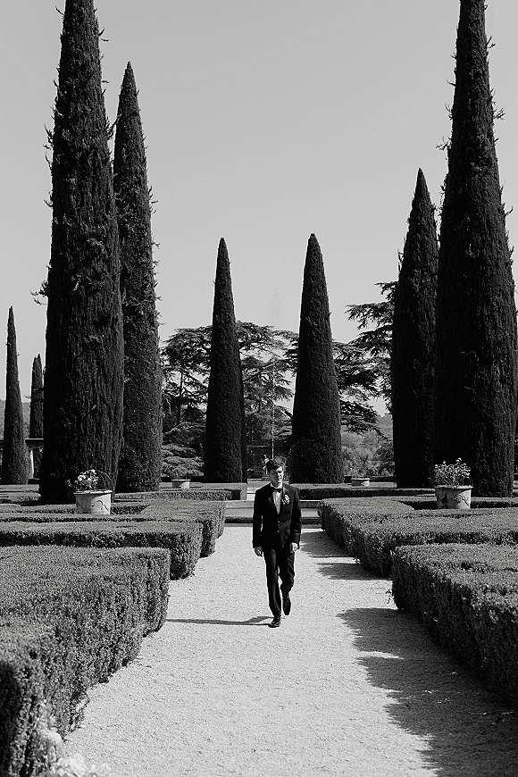 Groom portrait of a tuxedo groom in a black tuxedo with bow tie and boutonniere, walking a gravel path in a formal garden with cypress trees