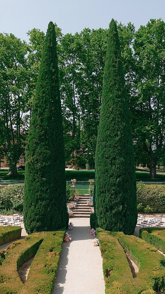 Ceremony setup for an outdoor garden ceremony with wedding aisle flowers, chair rows flanking stone steps in a formal cypress-lined garden