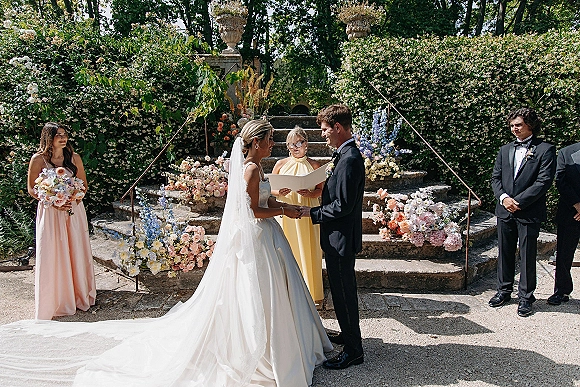 Wedding vows as bride and groom holding hands with bridal veil flowing, officiant reading beside floral-lined stone steps in a hedge garden