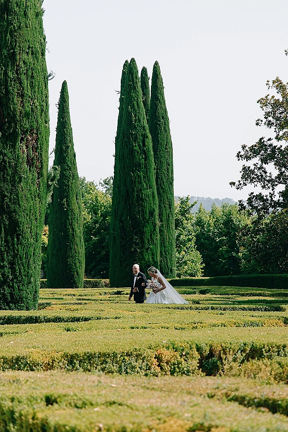 Bride walking portrait with her father, holding a pastel bouquet and long veil train along manicured garden hedges and cypress trees