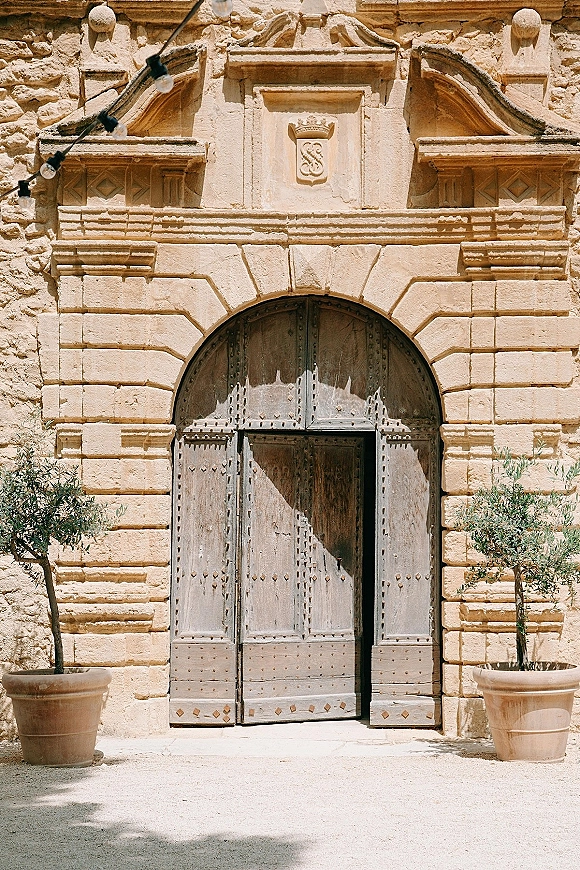 Venue entrance with arched wooden double doors and iron studs, flanked by potted trees in terracotta planters on a stone facade with string lights