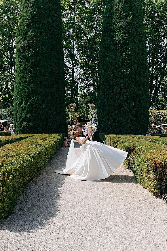 Wedding kiss portrait of groom dipping bride in veil and gown holding bouquet on a gravel path in a formal garden with guests behind