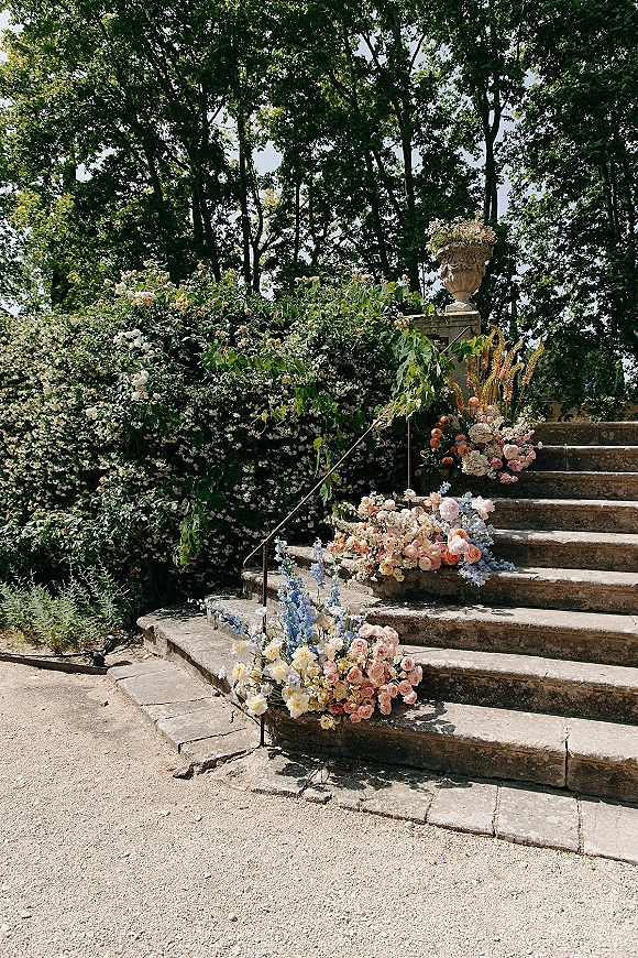 Ceremony aisle florals with roses, delphinium, and greenery lining stone steps and railing beside a gravel garden path