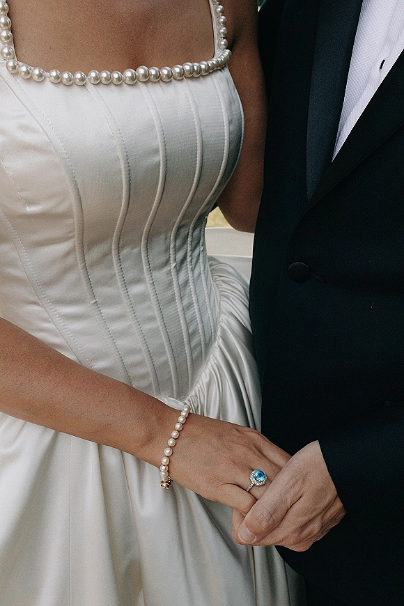 Bridal gown details with satin corset wedding dress, pearl necklace and bracelet beside groom’s tuxedo, highlighting an aquamarine ring on hands