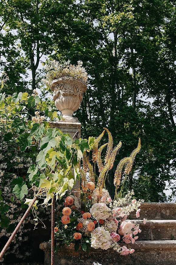 Wedding floral installation with pink and peach blooms and greenery vines in an urn planter cascading down stone steps on a garden terrace