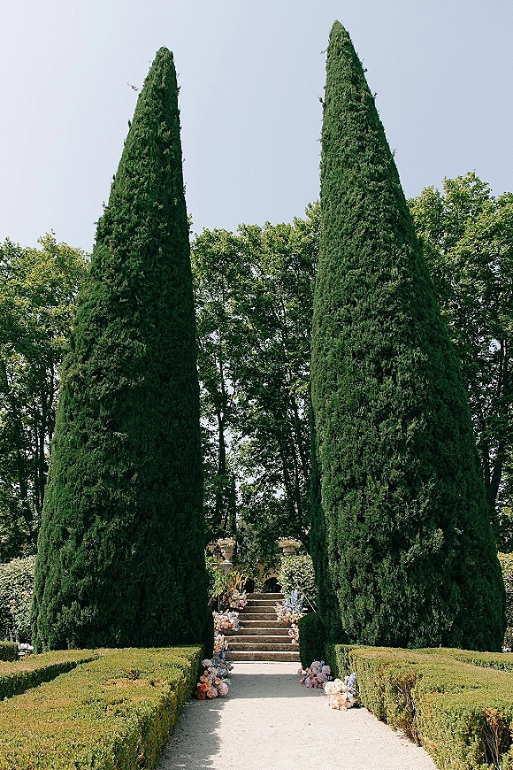 Ceremony aisle decor with pastel floral arrangements lining an outdoor ceremony aisle along a hedge-trimmed garden walkway to stone steps