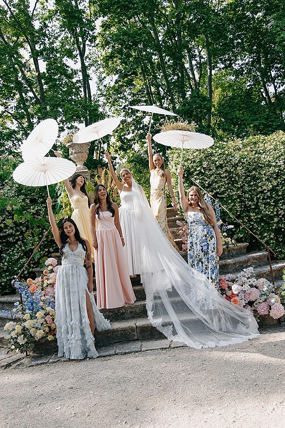 Bride with bridesmaids in a bridal party portrait holding white parasols, her long veil flowing on a stone garden staircase lined with florals