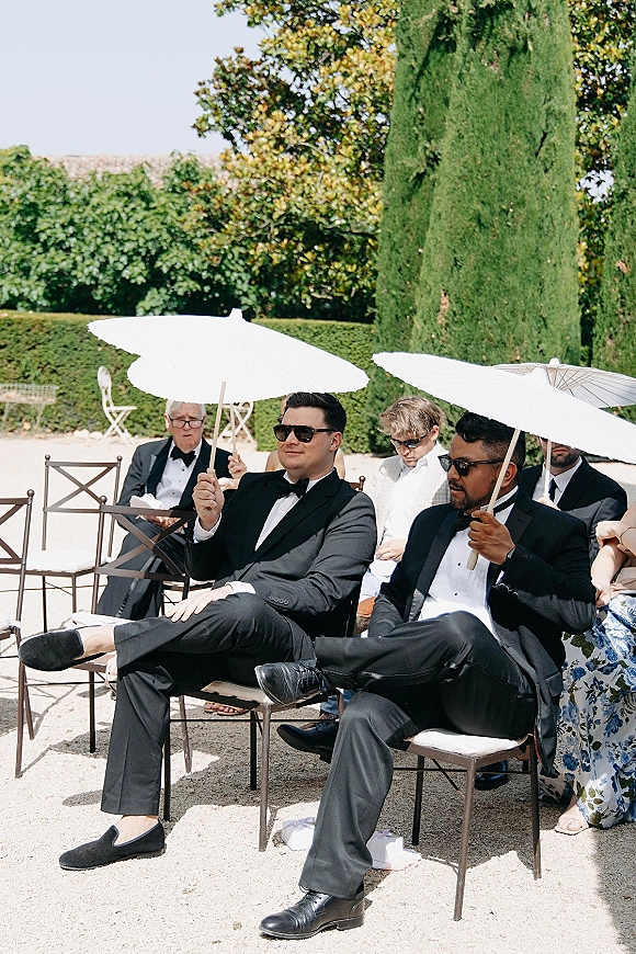 Wedding guests in black tuxedos sit in ceremony chairs wearing sunglasses, some holding white parasols in a garden courtyard with cypress trees