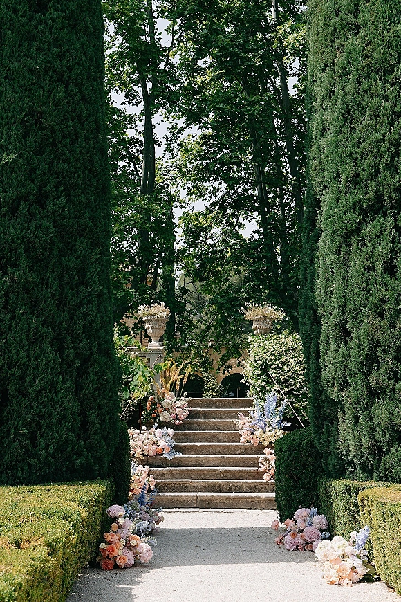 Ceremony aisle decor with garden wedding aisle floral clusters in urn planters along stone steps, trimmed hedges, and cypress trees