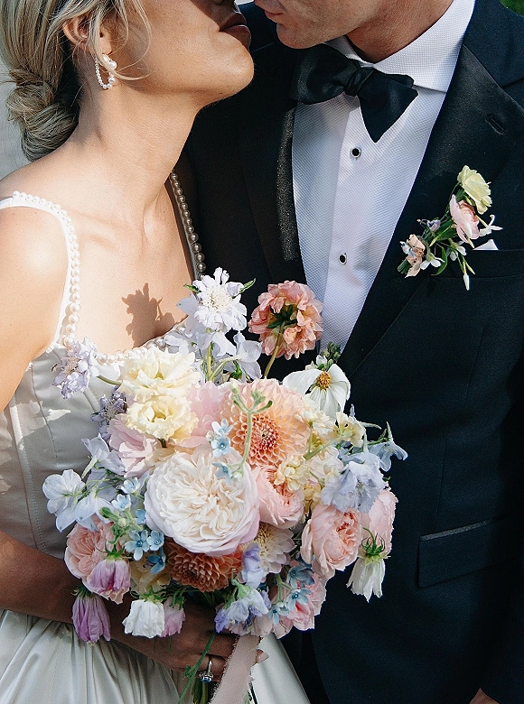 Wedding couple portrait of bride and groom close up sharing a nose kiss, her bouquet with ribbon wrap beside his tuxedo and bow tie on a neutral wall