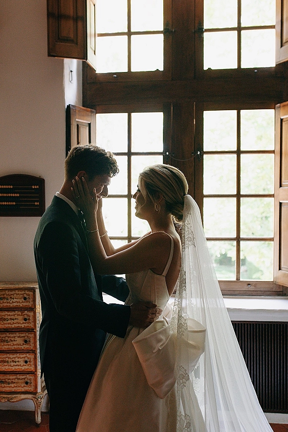 Couple portrait with bride and groom silhouette, bride touching groom’s face in window light beside a large wooden-framed window