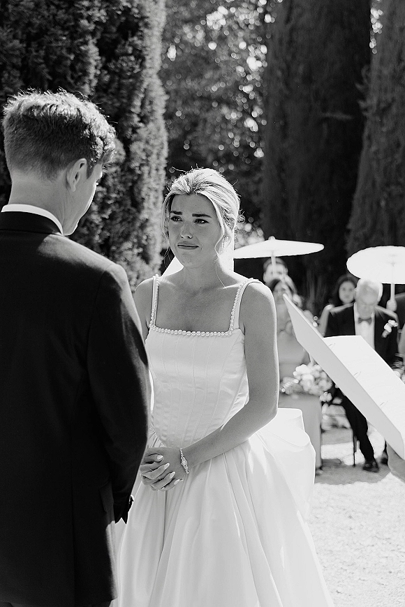 Wedding vows as bride in beaded strap gown holds groom’s hand during outdoor wedding ceremony, guests with parasols in sunlit garden