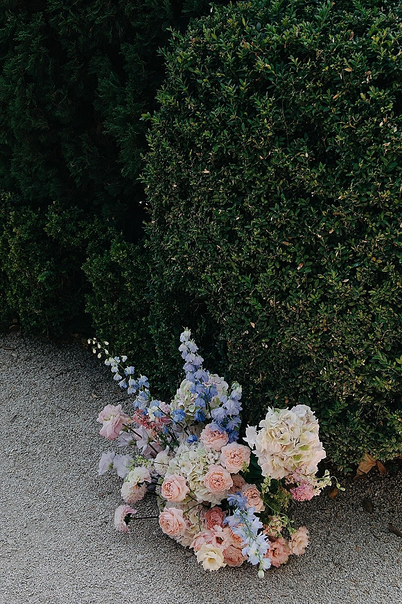 Wedding florals in a pastel wedding flowers arrangement with hydrangea, roses, and blue delphinium on gravel beside a green hedge