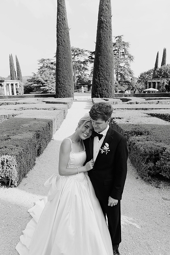Couple portrait of bride in a strapless ball gown leaning on groom in tuxedo with boutonniere on a gravel path in a formal garden