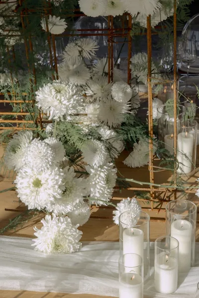Wedding tablescape with a white floral centerpiece, greenery, and pillar candles in glass cylinders on a wood tabletop with place settings