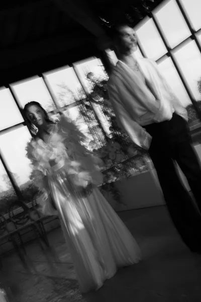 Couple portrait in black and white wedding portrait style, bride in tulle ruffle sleeves beside groom at large windows in an indoor venue