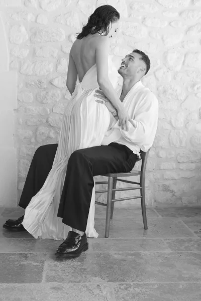 Couple portrait in a black and white wedding portrait, groom seated on a chair looking up while holding hands with bride in low back gown by a stone wall