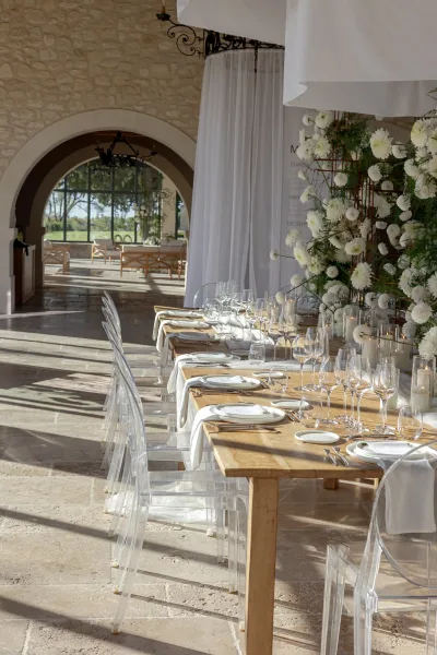 Reception tablescape with wood banquet table wedding details, ghost chairs, taper candles, and white florals beneath stone arches and chandeliers